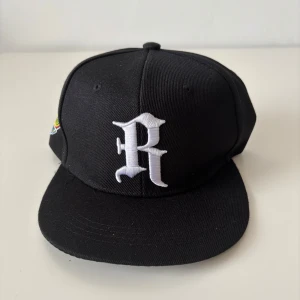 Svart Baseballkeps  - Snygg svart snapback keps med stor vit broderad R på fronten och färgglad broderad detalj på sidan. Justerbar baktill med klassisk plastspänne. Perfekt för dig som gillar streetwear och vill sticka ut med en cool accessoar. Helt ny.