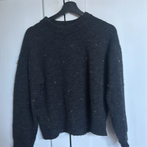 Mörkgrå stickad tröja med pärlor H&M - Mysig mörkgrå stickad tröja från H&M i storlek XS. Tröjan har ett enkelt, klassiskt snitt med ribbad hals och muddar. Små pärlor är diskret placerade över hela tröjan för en extra touch. Perfekt för dig som gillar stilrena detaljer.