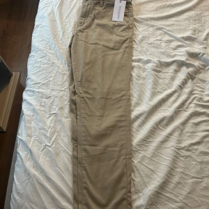 Beige chinos från Selected Homme - Snygga beige chinos från Selected Homme i modellen Marco. Byxorna har klassisk rak passform, bälteshällor och stängs med dragkedja och knapp. Perfekta för en clean och stilren look. Tillverkade i mjuk bomull för skön känsla hela dagen.