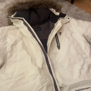 Vit dunjacka Peak Performance med fuskpäls - Snygg vit dunjacka från Peak Performance med avtagbar huva och fuskpäls. Jackan har svarta detaljer, dragkedja, flera fickor och är fodrad med svart insida. Perfekt för kalla dagar och har en klassisk, stilren look.