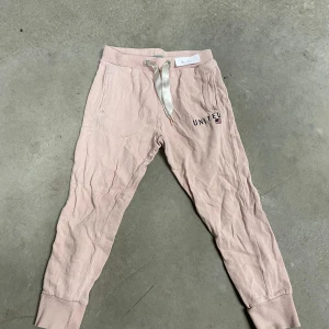 Rosa mjukisbyxor Iconic Sport 110 - Ljusrosa mjukisbyxor från Iconic Sport med broderad text 'UNITED' och liten flagga på ena benet. Byxorna har ribbade muddar, sidofickor och bred satinliknande snörning i midjan. Perfekta för chill dagar eller träning.