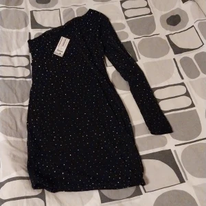 Svart glittrig one shoulder klänning New Yourker  - Supersnygg svart kortklänning från New Yourker med glittriga detaljer. Klänningen har en trendig one shoulder-design och långa ärmar. Perfekt för dig som vill sticka ut med en cool och elegant look. Materialet är mjukt och följsamt för en bekväm passform. Storlek:XL. Prislappen är kvar.