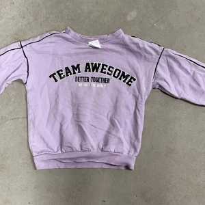 Ljuslila sweatshirt Team Awesome Uniq Studio - Ljuslila sweatshirt från Uniq Studio med svart text 'TEAM AWESOME' och 'BETTER TOGETHER' framtill. Tröjan har svarta detaljer längs axlarna, ribbade muddar och rund hals. Perfekt för dig som vill sticka ut med en cool statementtröja.