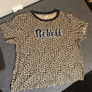 Leopardmönstrad t-shirt med text - Cool t-shirt med leopardmönster och svart texten 'Rebell' framtill. T-shirten har svart kant vid halsen och är i klassisk kortärmad modell. Perfekt för dig som vill sticka ut med en edgy vibe. 