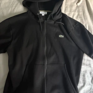 Svart hoodie från Lacoste med dragkedja - Svart hoodie från Lacoste i klassisk passform med dragkedja framtill och huva med snörning. Tröjan har två fickor och den ikoniska gröna krokodilloggan på bröstet. Perfekt för en chill och stilren look.