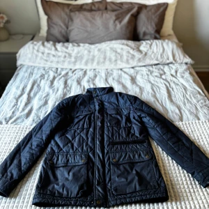 Mörkblå quiltad jacka med fickor - Snygg mörkblå quiltad jacka med hög krage och flera praktiska fickor framtill. Jackan har dragkedja och tryckknappar, samt ett diskret rutmönster i quiltningen. Perfekt för kyligare dagar och har en klassisk, stilren look. Skriv för fler bilder mvh Eddie