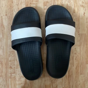 Svarta och vita Lacoste sandaler - Säljer ett par sandaler från Lacoste i svart och vitt med bred rem och guldigt krokodilmärke på sidan. de finns små defekter men inget som syns särskilt mycket. köpt på JD, ordinariepris är 600kr