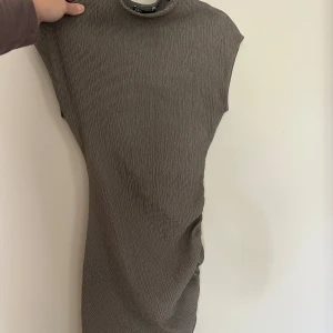 Brun kortärmad klänning från Zara - Säljer en brun kortklänning från Zara med diskret rutigt mönster och hög hals. Klänningen har en tight passform och är ärmlös, perfekt för en stilren look. Materialet är mjukt och stretchigt, vilket gör den bekväm att bära hela dagen.