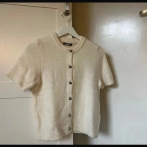 Beige kortärmad kofta med knappar - Superfin beige kofta med korta ärmar och rund hals. Koftan har snygga knappar framtill och är stickad i ett mjukt material som känns lyxigt. Perfekt att slänga över en topp för en chill men ändå stilsäker look. Något nopprig, passar mer S