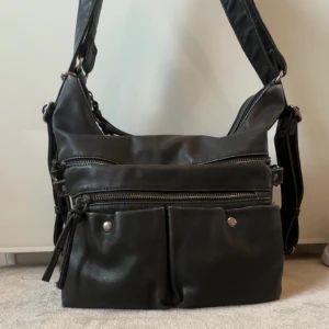 Oandvänd axelväska från Dressy. Original pris: 400-500 - Snygg svart axelväska i skinn med flera dragkedjor och praktiska ytterfickor framtill. Väskan har bred axelrem och silverfärgade detaljer som ger en cool vibe. Perfekt storlek för att få plats med allt du behöver till vardags.