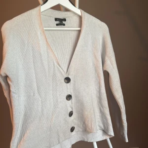 Vit kofta från Massimo Dutti - Ribbstickad beige kofta från Massimo Dutti med v-ringning och fyra stora knappar framtill. Koftan har lång ärm och en något croppad passform, perfekt att slänga över en t-shirt eller linne. Enkel och stilren design som funkar året runt.🍂🍁