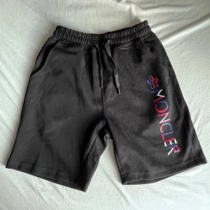 Svarta Moncler shorts med tryck - Svarta shorts från Moncler med elastisk midja och snörning. Snyggt Moncler-tryck i rött, blått och vitt längs ena benet. Perfekta för chill dagar eller när du vill ha en clean streetstyle-look.