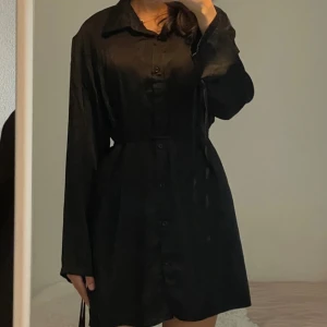 Svart skjortklänning med knappar - Stilren svart skjortklänning med lång ärm och knappar hela vägen framtill. Klänningen har en klassisk krage och markerad midja, vilket ger en snygg siluett. Perfekt för dig som gillar minimalistisk och tidlös stil.