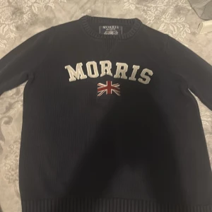 Mörkblå tröja från Morris XS - Mörkblå tröja från Morris i storlek XS med broderad logga och brittisk flagga på bröstet. Klassisk rund halsringning och ribbade muddar. Perfekt för dig som gillar preppy stil och snygga detaljer.