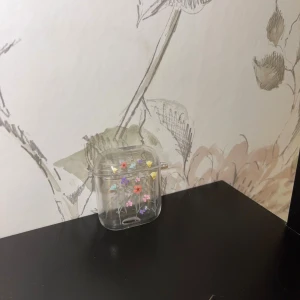 Transparent blommig AirPods-fodral - Säljer ett genomskinligt AirPods-fodral med söta små blommor i olika färger som dekoration. Fodralet är gjort i plast och har ett smidigt lock som skyddar dina hörlurar. Perfekt accessoar för dig som vill ha något gulligt och personligt till dina AirPods.