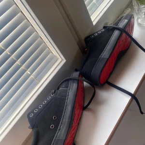 Svarta sneakers med röd sula - Snygga svarta sneakers från Christian Louboutin med ikonisk röd sula och svarta snören. Ovandelen är i mocka och insidan har en mjuk, ljusbrun läderdetalj. Skorna har rund tå och klassisk sneakerlook med metallöljetter.