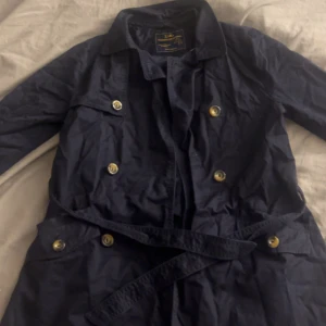 Mörkblå trenchcoat från Zara - Snygg mörkblå trenchcoat från Zara med guldiga knappar och bälte i midjan. Jackan är vattenavvisande och har klassisk krage samt långa ärmar och har dubbla knapprader framtill. Säljer pga den är tyvärr för liten.
