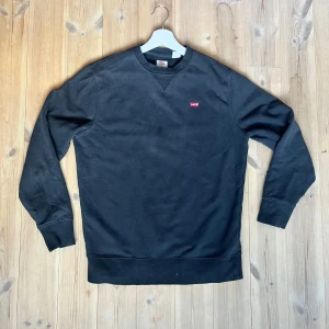 Svart sweatshirt från Levi's - En stilren svart sweatshirt från Levi's med liten röd logotyp på bröstet. Tröjan har rund halsringning, långa ärmar och ribbade muddar vid ärmslut och nederkant. Perfekt för en avslappnad look och enkel att matcha med jeans eller andra byxor.
