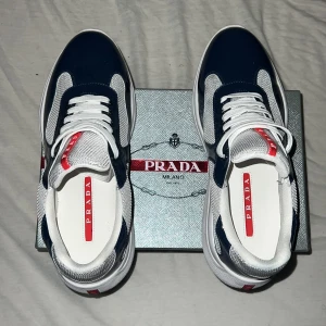 Prada sneakers i marinblå och vitt - Snygga Prada sneakers i marinblått och vitt med röda detaljer och tydlig logga på plösen och hälen. Ovandelen är i mesh och glansigt syntetmaterial, med vita snören och en chunky vit sula. Perfekta för dig som vill ha en exklusiv och sportig look. Pris går att diskuteras!