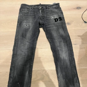 Svarta Dsquared2 jeans med D2-tryck - Snygga svarta jeans från Dsquared2 med D2-tryck på ena fickan. Jeansen har en tvättad look med slitningar och små färgstänk för en cool vibe. Tillverkade i bomull med lite elastan för skön stretch. Klassisk femficksmodell och raka ben.