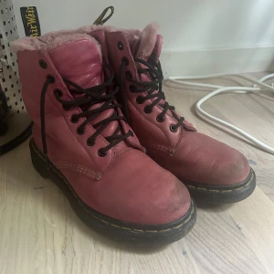 Rosa Dr. Martens boots med foder - Säljer ett par rosa Dr. Martens boots med svart snörning och mjukt rosa foder. Skorna har klassisk gul söm runt sulan och är tillverkade i skinn. Perfekta för kalla dagar med sin varma insida och robusta design.
