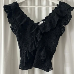 Svart stickad volangblus från Zara - Supersnygg svart blus från Zara med volanger längs v-ringningen och axlarna. Blusen är stickad med spetsdetaljer och har en croppad passform. Perfekt för dig som vill ha en trendig och unik topp med mycket detaljer.