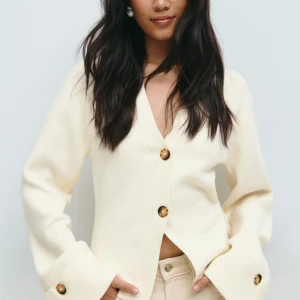 Krämvit kofta från Glamroot med knappar - Stilren krämvit kofta från Gina Tricot med v-ringning och stora bruna knappar framtill och vid ärmsluten. Koftan har lång ärm, rak passform och är tillverkad i mjukt stickat material. Perfekt för lager-på-lager och enkel att matcha. Helt slutsåld, aldrig använd.