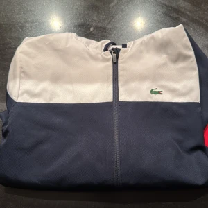 Lacoste Sport vindjacka i rött, blått och vitt - Snygg vindjacka från Lacoste Sport i färgblock med vitt upptill, marinblått i mitten och rött nedtill. Jackan har hel dragkedja, huva och klassisk Lacoste-logga på bröstet. Helt nytt skick, pris kan diskuteras
