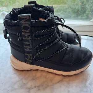Svarta vinterboots från The North Face - Säljer ett par svarta vinterboots från The North Face i storlek 38. Boots med snörning, tjock vit sula och fuskpälsfoder. Yttermaterial i syntet och textil, med stora logoband på sidan och reflexdetaljer på snörena. Perfekta vinterskor! Knappt använda 