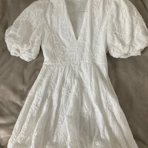 Vit kortklänning med broderade blommor - Superfin vit kortklänning med puffärmar och djup v-ringning. Klänningen har stora broderade blommor över hela tyget och är sydd i ett lätt material som känns luftigt. Perfekt för sommarens alla tillfällen och ger en romantisk vibe.