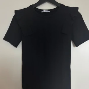 Svart ribbad topp med volang från Zara!💗 - Svart ribbad topp från Zara med korta ärmar och snygga volangdetaljer på axlarna. Toppen har rund halsringning och är tillverkad i ett stretchigt material som sitter skönt på kroppen. Endast använd ett fåtal gånger. 💗