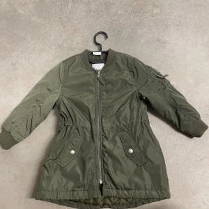 Olivgrön bomberjacka från Lindex Kids - Snygg olivgrön bomberjacka från Lindex Kids med ribbade muddar, dragkedja framtill och cool ficka med dragkedja på ärmen. Jackan har två fickor med tryckknappar och markerad midja för en schysst passform. Perfekt för dig som gillar streetstyle.