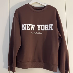 Brun sweatshirt med New York-tryck - Brun sweatshirt i storlek M med vitt New York-tryck på bröstet. Tröjan har rund halsringning, ribbade muddar vid ärmslut och nederkant samt en avslappnad passform. Perfekt för dig som gillar streetstyle och vill ha en enkel men snygg look.