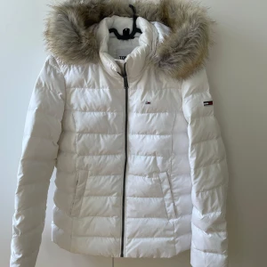 Vit vinterjacka från Tommy Hilfiger  - Snygg vit vinterjacka från Tommy Hilfiger med avtagbar huva och fuskpäls. Jackan har dragkedja framtill, två fickor och diskret logga på bröstet och ärmen. Perfekt för kalla dagar och ger en clean look med sin quiltade design. Köpt på hede fashion, endast använt en gång.