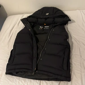 Svart dunväst från Everest med huva - Svart dunväst från Everest i storlek 36. Västen har en stor, vadderad huva, dragkedja framtill och tryckknappar. Den är quiltad och har två sidofickor. Perfekt för kyliga dagar när du vill ha en sportig och clean look.