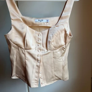 Champagnefärgad satin korsett från NA-KD - Supersnygg champagnefärgad korsett från NA-KD i glansig satin. Toppen har breda axelband, markerade kupor och dekorativa knappar framtill. Figurnära passform som ger en riktigt trendig siluett. Perfekt att styla med jeans eller kjol för en chic look. Helt ny utan prislapp.