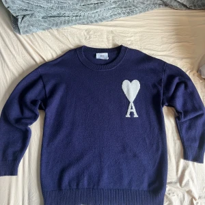 Mörkblå stickad tröja från Ami - Säljer en mörkblå stickad tröja från Ami med rund halsringning och ribbade muddar. På bröstet finns ett stort vitt hjärta med bokstaven A. Perfekt för dig som gillar stilrena och trendiga plagg. Storlek M