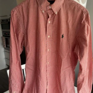 Rutig röd skjorta från Ralph Lauren - Säljer en snygg röd och vit rutig skjorta från Ralph Lauren. Skjortan har lång ärm, klassisk krage med knappar och broderad logga på bröstet. Tillverkad i stretchig bomull för en bekväm slim fit-passform.