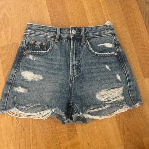 Högmidjade jeansshorts Stradivarius - Snygga blå högmidjade jeansshorts från Stradivarius med slitna detaljer och fransig kant nertill. Klassisk femficksmodell med knapp och dragkedja. Perfekta för en chill sommarlook.