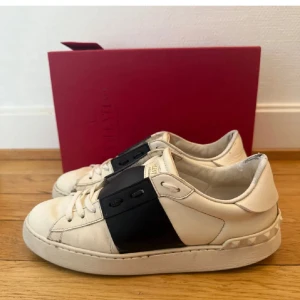 Valentino Garavani Open sneakers vit/svart - Snygga Valentino Garavani Open sneakers i vitt skinn med bred svart läderdetalj över snörningen. Klassisk rund tå, platt sula och ikoniska nitar baktill. Kommer med box. 