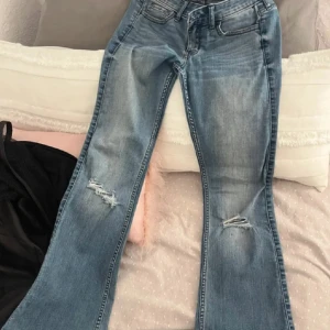 Blåa Hollister Flare Jeans W25 - Snygga blåa flare jeans från Hollister med slitningar på knäna. Jeansen har klassisk femficksdesign, låg midja och utsvängda ben. Perfekta för dig som gillar en trendig och avslappnad look.