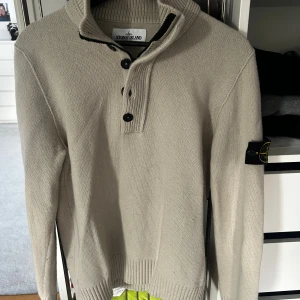 Beige stickad tröja från Stone Island - Säljer en beige stickad tröja från Stone Island med hög krage och tre knappar framtill. Tröjan har lång ärm och den klassiska Stone Island-loggan på vänster ärm. Perfekt för dig som gillar stilren och enkel design. Äkta såklart