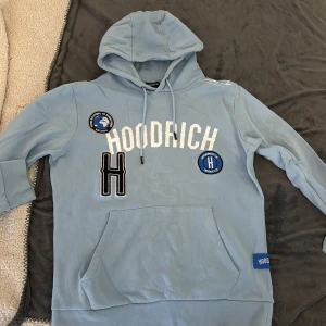 Ljusblå hoodie från Hoodrich M - Ljusblå hoodie från Hoodrich med broderade patches och stor vit logga framtill. Tröjan har huva med snörning, känguruficka och ribbade muddar. Snygga blå och svarta detaljer samt tryck på axeln. Perfekt för en chill streetwear-look.