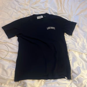 Mörkblå t-shirt från Les Deux - Säljer en svart t-shirt från Les Deux med vit broderad logga på bröstet. Klassisk passform med rund hals och korta ärmar. Skön och stilren, perfekt till jeans eller shorts. Materialet är mjuk bomull som känns nice mot huden.