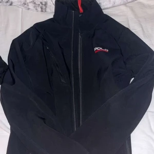Svart vindjacka från Polo - Svart vindjacka från Polo med hög krage och diskret röd detalj på insidan. Jackan har dragkedja framtill och en bröstficka med dragkedja. Polo-loggan är broderad i vitt och rött på vänster bröst. Perfekt för dig som gillar stilren och sportig look.