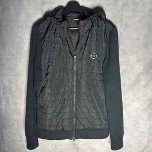 Svart hybridjacka från J.Lindeberg - Snygg svart hybridjacka från J.Lindeberg med quiltad front och huva. Ärmarna är stickade och ger en cool kontrast till det glansiga materialet på kroppen. Jackan har dragkedja framtill, sidofickor och en liten bröstficka med logga. Båda fickorna går ej att dra igen helt men ska gå att fixa.