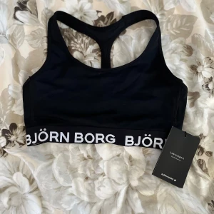 Svart sporttopp från Björn Borg XS  - Sporttopp från Björn Borg i svart med bred resår nertill där loggan är tryckt i vitt. Modellen har brottarrygg och är tillverkad i ett stretchigt syntetmaterial, perfekt för träning. Låg support och stilren design.