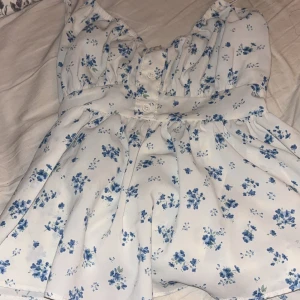 Vit blommig volang topp med rosett på ryggen - Supersöt vit topp med blått blommönster och knappar framtill. Klänningen har smala axelband, volangkant och en stor rosett i ryggen som ger en öppen rygg. Perfekt för varma dagar och har en luftig, flowy passform.