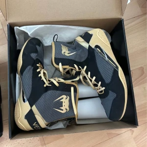 Svarta och beige Venum boxningsskor - Säljer ett par svarta och beige boxningsskor från Venum med högt skaft och snörning. Skorna har meshpaneler för ventilation och en platt sula för bra grepp. Stora Venum-loggor på sidorna och cool sportig design.