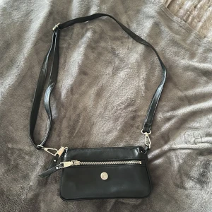 Svart axelväska i skinn - märket ”Ulrika” - Snygg svart axelväska i skinn från Ulrika med justerbar rem, silverfärgade detaljer och dragkedja framtill. Väskan har flera fack med blommigt innerfoder och en stilren rund logga i metall på framsidan. Perfekt storlek för det viktigaste du behöver ha med dig. Nypris: 499 kr